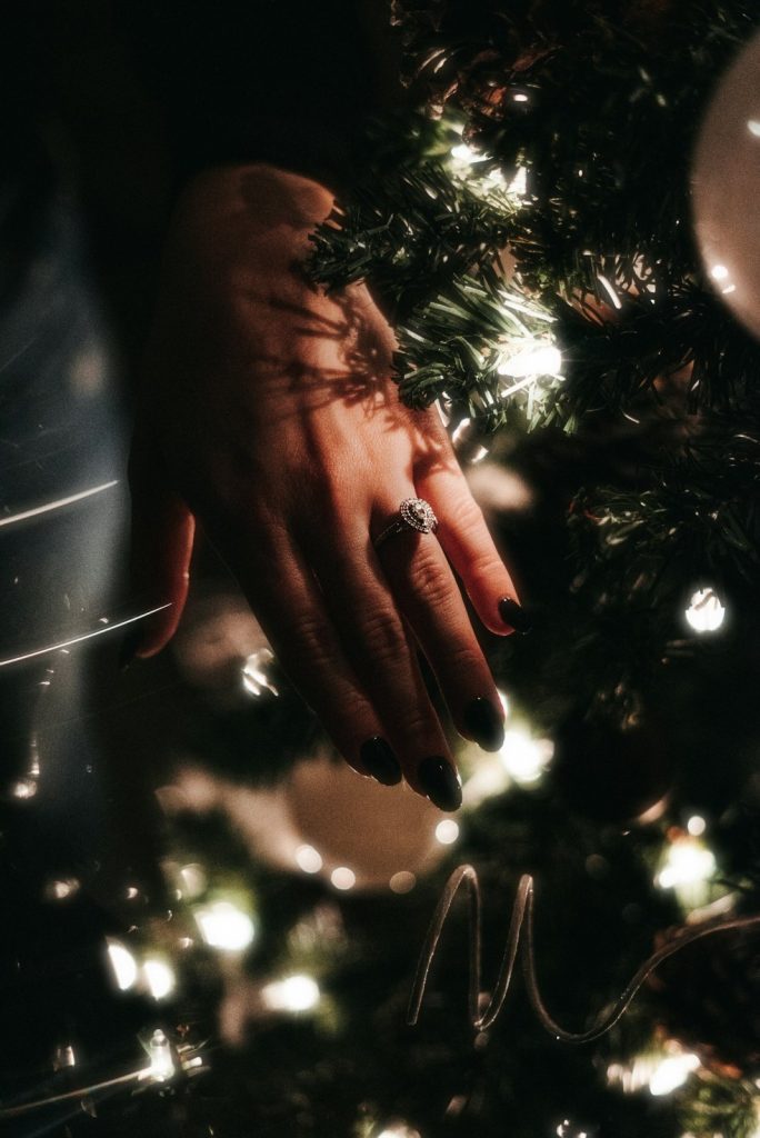 A hand wearing a sparkling engagement ring against the lights of a Christmas tree after a Christmas proposal